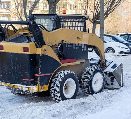 Ein gelber Schneepflug räumt Schnee in einem Parkplatz, hinter dem Autos stehen. Ein Gebäude ist im Hintergrund sichtbar.