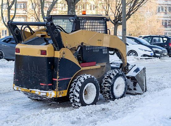 Ein gelber Schneepflug räumt Schnee in einem Parkplatz, hinter dem Autos stehen. Ein Gebäude ist im Hintergrund sichtbar.