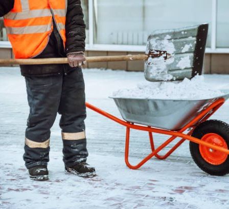 Ein Mann in einer Warnweste schaufelt Schnee in einen Schubkarren. Der schneebedeckte Boden und ein Gebäude mit Glasfenstern sind im Hintergrund.