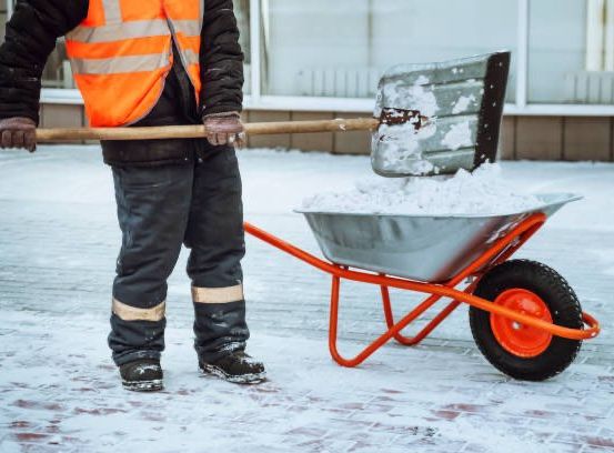 Ein Mann in einer Warnweste schaufelt Schnee in einen Schubkarren. Der schneebedeckte Boden und ein Gebäude mit Glasfenstern sind im Hintergrund.