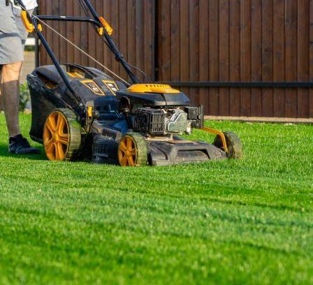 Eine Person in Shorts mäht einen gepflegten Rasen mit einem modernen, gelben und schwarzen Rasenmäher und steht vor einem Holzzaun.