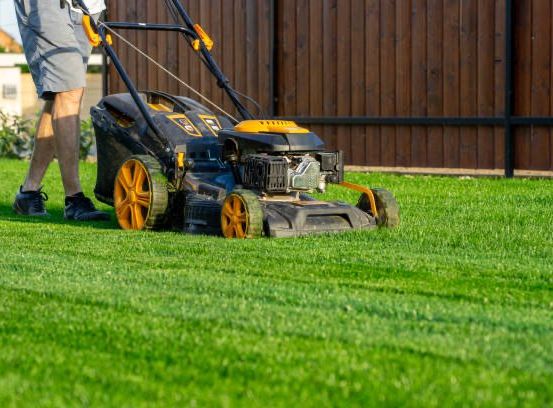Eine Person in Shorts mäht einen gepflegten Rasen mit einem modernen, gelben und schwarzen Rasenmäher und steht vor einem Holzzaun.