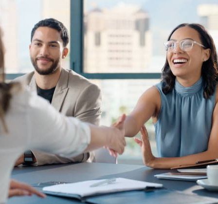 Drei Personen, zwei Männer und eine Frau, sitzen um einen Tisch herum und führen ein Gespräch. Die Frau mit Brille lächelt und hält einen Stift. Der Mann neben ihr hält ihre Hand, und der andere Mann lächelt. Sie sind von Büromaterialien umgeben.