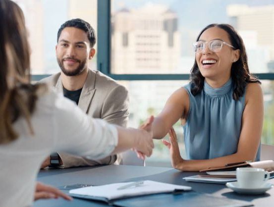 Drei Personen, zwei Männer und eine Frau, sitzen um einen Tisch herum und führen ein Gespräch. Die Frau mit Brille lächelt und hält einen Stift. Der Mann neben ihr hält ihre Hand, und der andere Mann lächelt. Sie sind von Büromaterialien umgeben.
