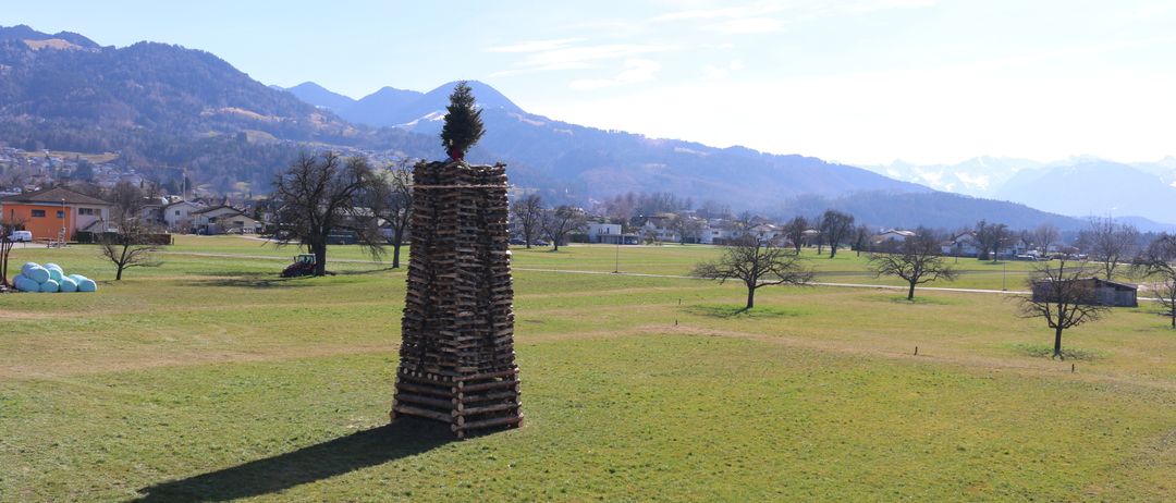 Ein hoher Stapel aus Holzscheiten mit einem kleinen Weihnachtsbaum darauf in einer weiten, grasigen Wiese mit Bergen im Hintergrund.