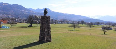 Ein hoher Stapel aus Holzscheiten mit einem kleinen Weihnachtsbaum darauf in einer weiten, grasigen Wiese mit Bergen im Hintergrund.
