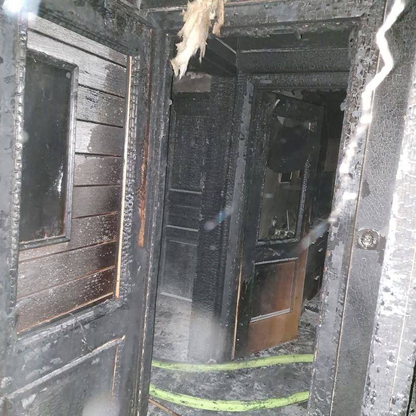 Ein verkohlter Innenraum eines Hauses zeigt zwei Türen, eine offene, die zu einem Flur mit Rauch und einem sichtbaren Schlauch führt.