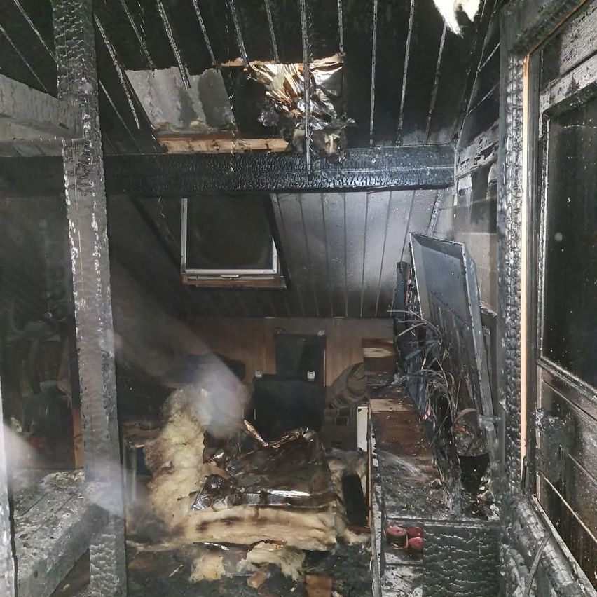 Ein verbrannter Raum mit beschädigter Decke, verstreuten Gegenständen und einem kaputten Monitor. Der Rauch und Ruß sind im ganzen Bereich sichtbar.