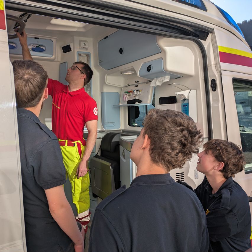 Ein Mann in roter Uniform zeigt einer Gruppe von Personen das Innere eines Krankenwagens.