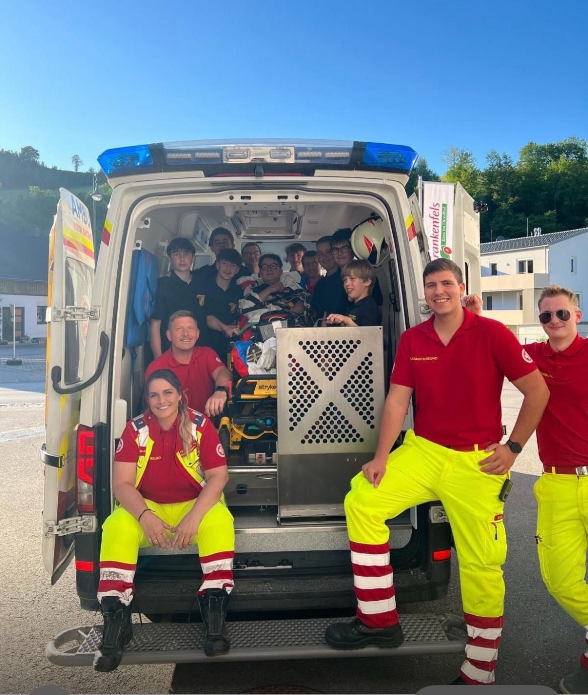 Eine Gruppe von Personen in roten und gelben Uniformen steht neben einem Krankenwagen. Eine Person sitzt auf einer Trage im Inneren. Eine andere Person hält einen Helm. Hinter ihnen stehen Gebäude und Bäume.