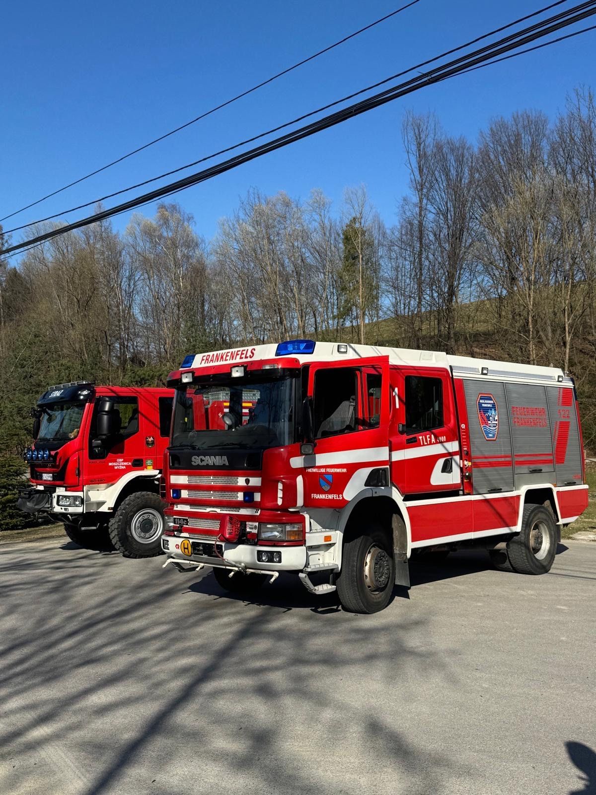 Zwei rote und weiße Scania-Löschfahrzeuge sind auf einem Parkplatz geparkt, mit einem bewaldeten Hügel im Hintergrund.