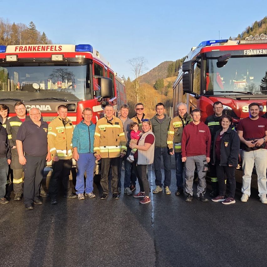 Eine Gruppe Feuerwehrleute und andere posieren vor zwei roten Feuerwehrwagen auf einer Straße mit Bergen im Hintergrund.