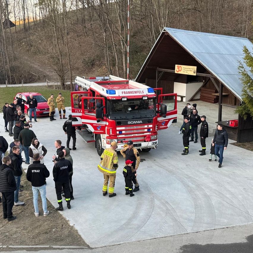 Ein roter Feuerwehrwagen mit der Aufschrift Frankenfels steht neben einer Hütte mit Dach und Schild. Mehrere Menschen in Uniformen und Jacken sind um den Wagen versammelt. In der Nähe steht ein rotes Auto, und weitere Menschen stehen oder gehen herum.