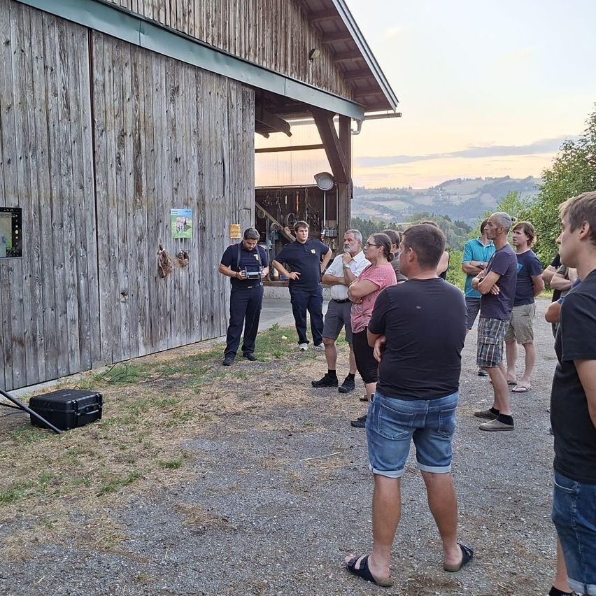 Eine Gruppe von Menschen, einschließlich Feuerwehrleuten, versammelt sich bei Sonnenuntergang vor einem Scheunengebäude. Ein Feuerwehrmann hält ein Gerät, während andere aufmerksam zuhören.