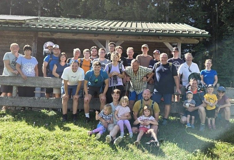Eine Gruppe von Menschen, einschließlich Erwachsener und Kindern, posiert für ein Foto vor einer Holzstruktur mit Metalldach. Sie stehen und sitzen auf einem Holzzaun und auf einem Rasen.