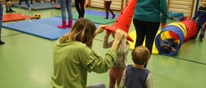 In einem Fitnessstudio hilft eine Frau einem Kind mit einem roten Kegel, während andere in der Nähe spielen. Blaue Matten und verschiedene Spielzeuge sind herum verstreut.