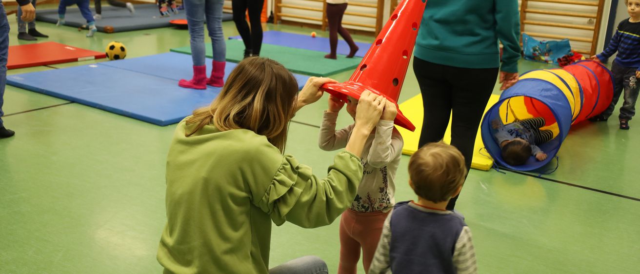 In einem Fitnessstudio hilft eine Frau einem Kind mit einem roten Kegel, während andere in der Nähe spielen. Blaue Matten und verschiedene Spielzeuge sind herum verstreut.