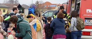 Familien mit Kindern und Erwachsene stehen auf Gras in der Nähe eines Feuerwehrwagens, einige halten Kinder. Ein Feuerwehrmann steht in der Nähe und interagiert mit Kindern. Gebäude mit Solarmodulen sind im Hintergrund.
