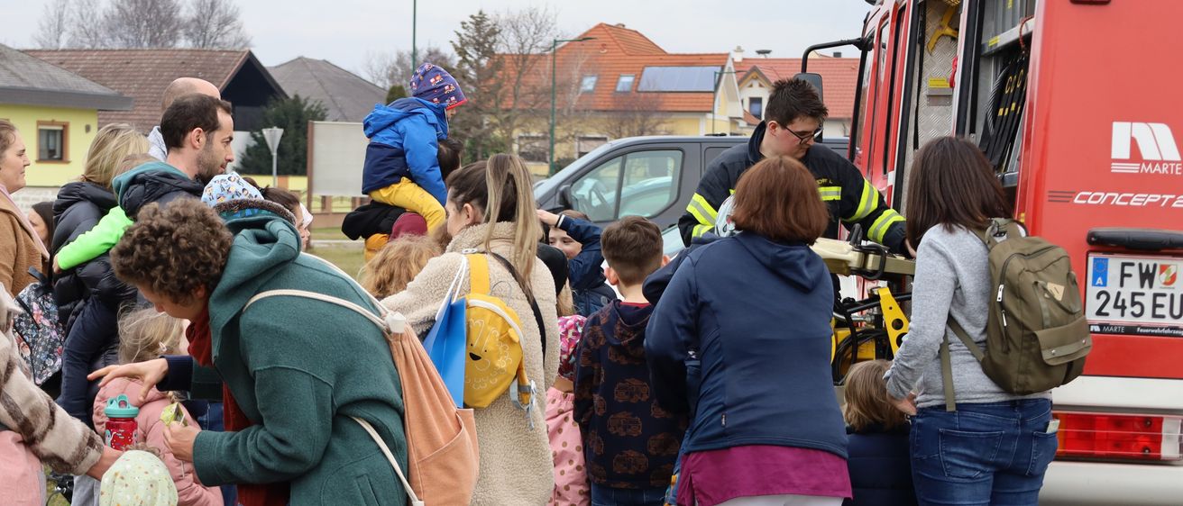 Familien mit Kindern und Erwachsene stehen auf Gras in der Nähe eines Feuerwehrwagens, einige halten Kinder. Ein Feuerwehrmann steht in der Nähe und interagiert mit Kindern. Gebäude mit Solarmodulen sind im Hintergrund.
