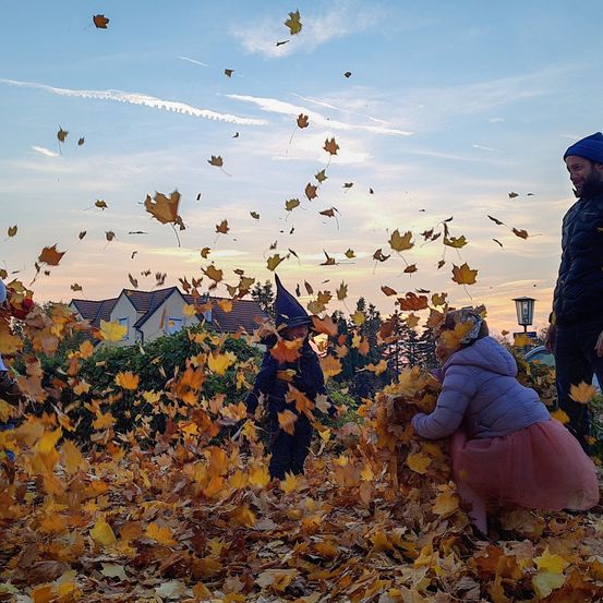 Eine Gruppe von Menschen hat Spaß mit Herbstblättern in einem Garten, mit Häusern im Hintergrund.