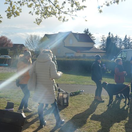 Eine Gruppe von Menschen, darunter Kinder und Erwachsene, versammelt sich in einem Park mit einem Haus im Hintergrund. Eine Person hält einen schwarzen Hund an der Leine.