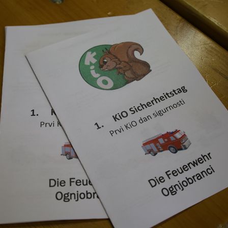 Zwei weiße Papiere mit 'Kio'-Logo, einem Eichhörnchen und Text über 'KIO Sicherheitstag', 'Privi KiO' und 'Feuerwehr'. Die Papiere haben Texte und Bilder zum Sicherheitstag und Feuerwehrleuten.