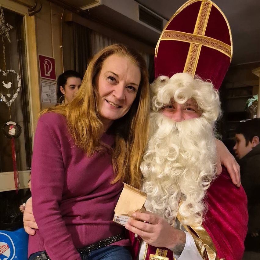 Eine Frau mit blonden Haaren lächelt und posiert für ein Foto mit einer Person in einem Weihnachtsmannkostüm. Sie halten einen Papierbeutel. Dahinter steht eine Person, und ein Feuerlöscher ist an der Wand montiert.