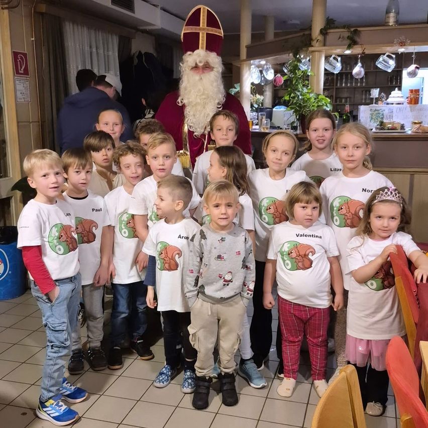 Eine Gruppe von Kindern in weißen T-Shirts mit Eichhörnchengrafiken steht vor einem als Weihnachtsmann verkleideten Mann, der von weiteren Kindern umgeben ist. Dahinter befindet sich eine Küche mit Regalen und einem Tisch mit Tassen und Flaschen.
