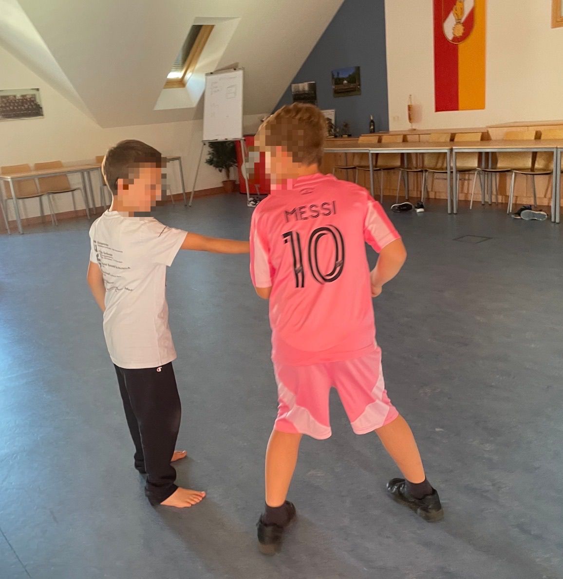 Zwei Kinder stehen sich in einem Raum gegenüber, eines trägt ein Messi-Trikot mit der Nummer 10. Sie sind barfuß.