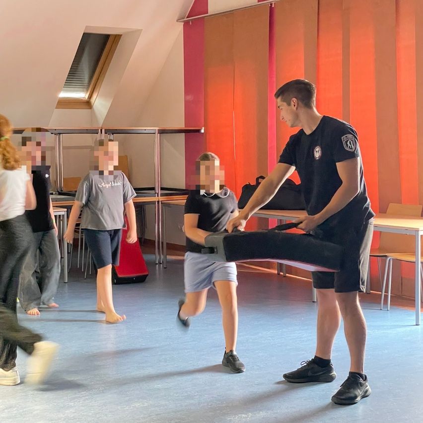 Eine Gruppe von Menschen in einem Fitnessstudio, ein Mann hilft einem Kind beim Kicken, andere laufen und stehen herum.