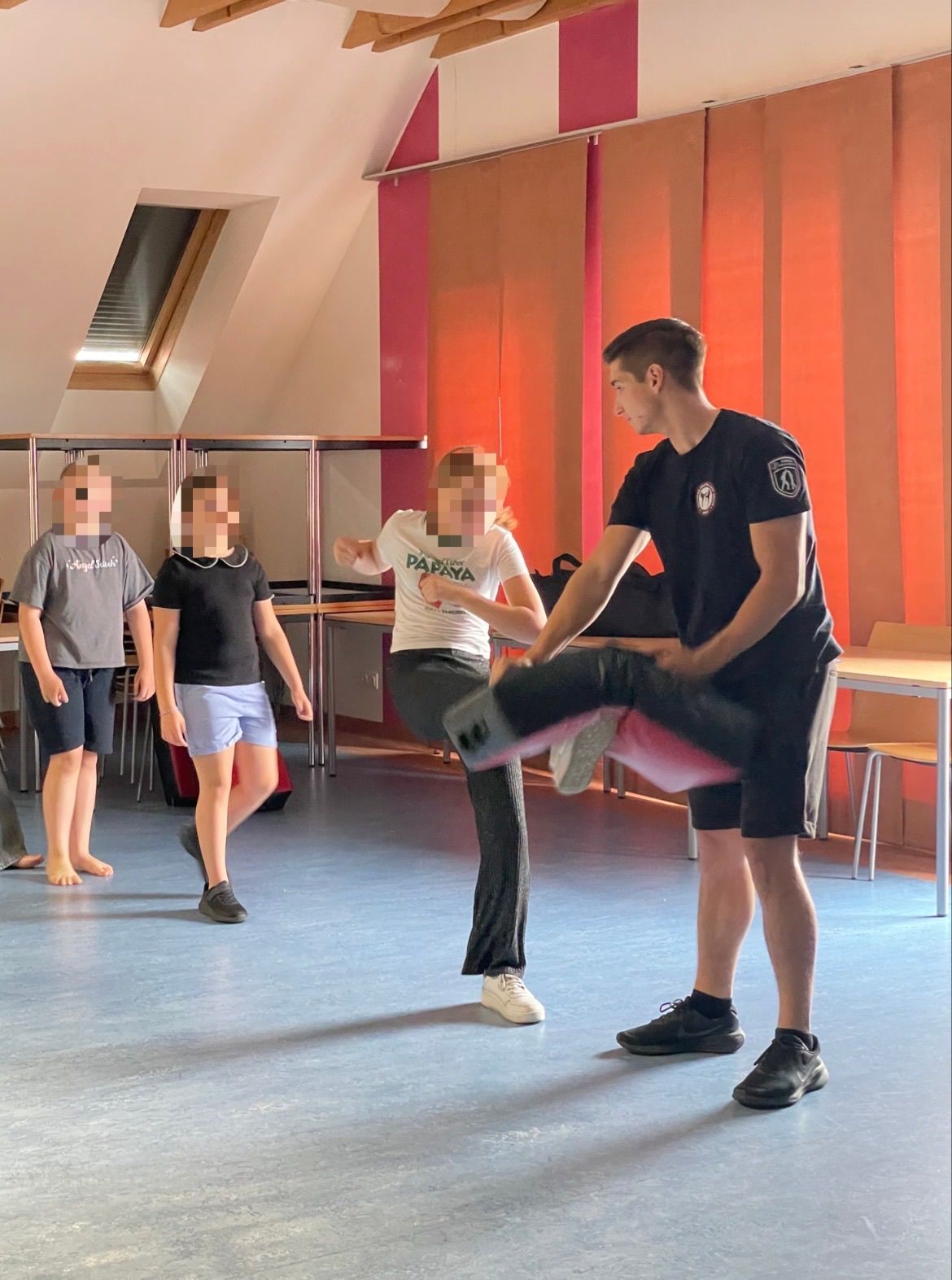 In einem Studio hilft ein Mann einer Frau in einer Kick-Position, während zwei andere zusehen. Dahinter sind ein Schreibtisch und Stühle aufgestellt.