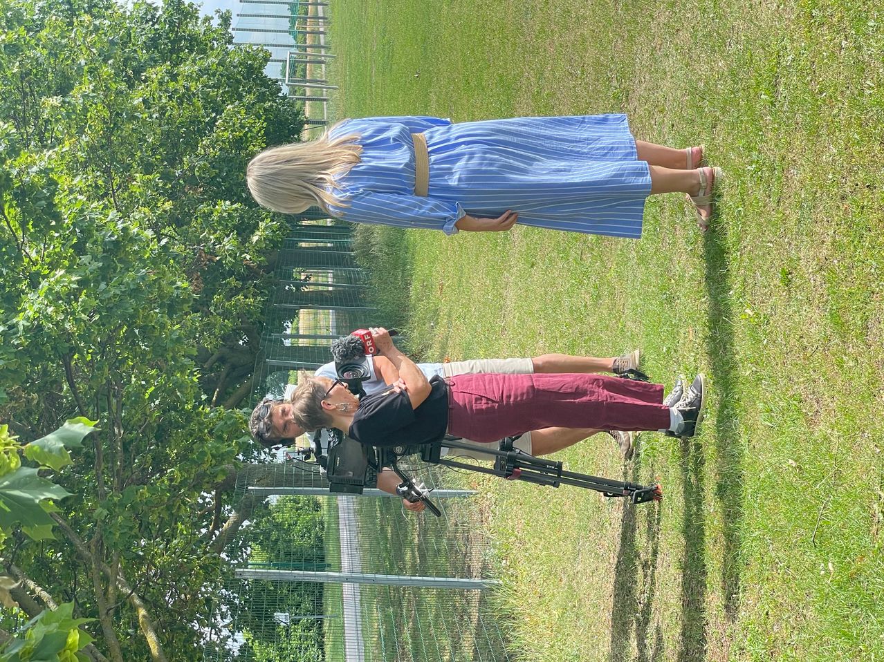 Zwei Personen filmen mit einer Kamera auf einem Stativ auf einer Grasfläche. Eine Frau liegt auf dem Gras, während die andere eine Kamera hält.