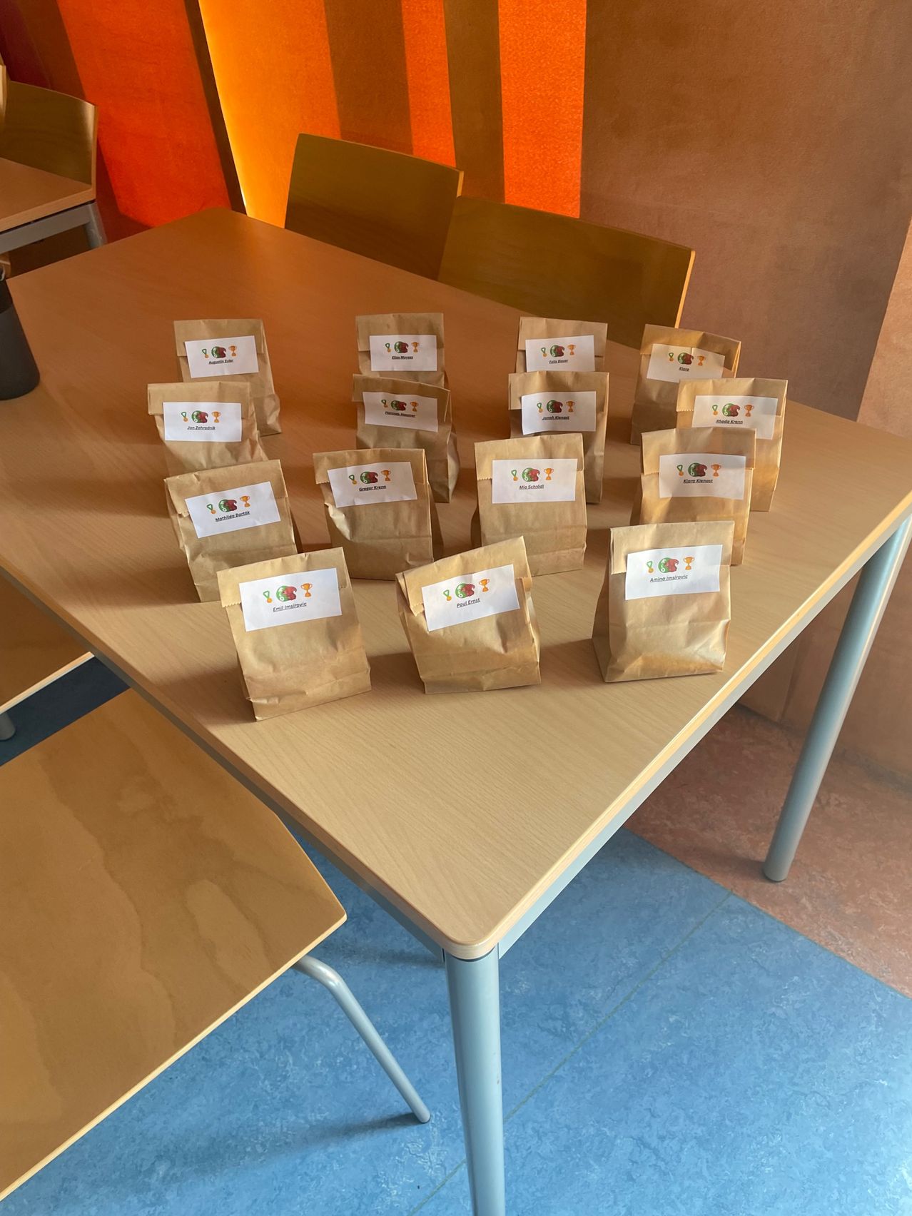 Ein Tisch mit braunen Papiertüten, jeweils mit einem weißen Etikett, ist in einer Reihe angeordnet. Der Tisch ist hellbraun und der Boden blau.