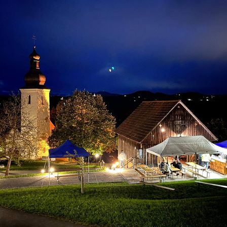Eine Dorfkirche mit Turm ist nachts beleuchtet. Daneben steht ein Bauernhaus mit Zelt und einer Versammlung von Menschen.