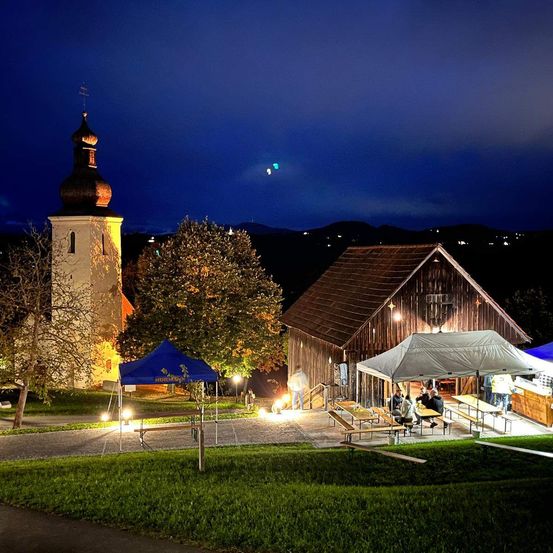 Eine Dorfkirche mit Turm ist nachts beleuchtet. Daneben steht ein Bauernhaus mit Zelt und einer Versammlung von Menschen.