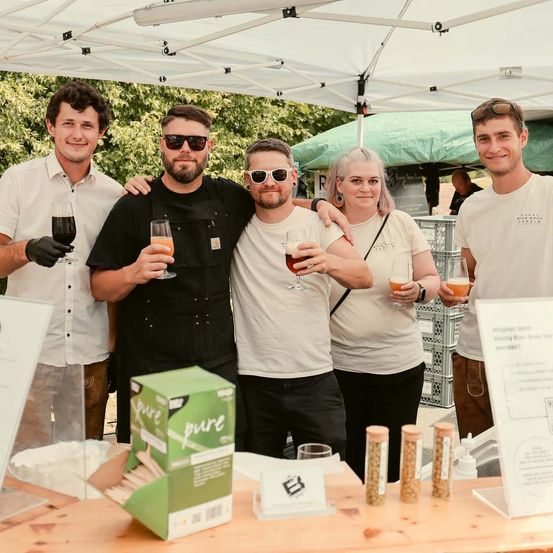 Vier Menschen stehen lächelnd unter einem Zelt und halten Weingläser in den Händen. Vor ihnen steht ein Tisch mit einer grünen Box, die 'pure' beschriftet ist. Dahinter ist ein grünes Zelt und einige Kisten sichtbar.