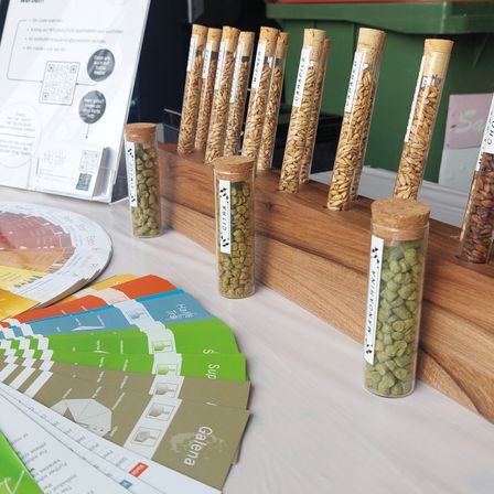 Auf einem Tisch sind verschiedene beschriftete Teströhrchen mit unterschiedlichen Körnern neben einem Farbkreis und Broschüren zu sehen.