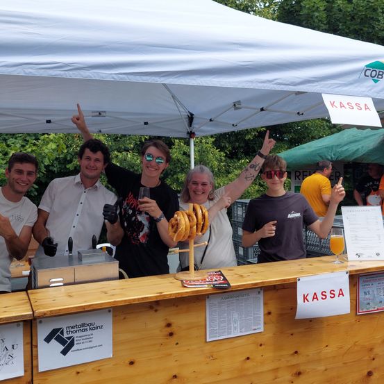 Eine Gruppe von Menschen posiert für ein Foto unter einem Zelt mit einem Schild, das 'Kassa' sagt. Sie stehen hinter einem Holztresen mit Brezeln, einem Glas Saft und einer Speisekarte.