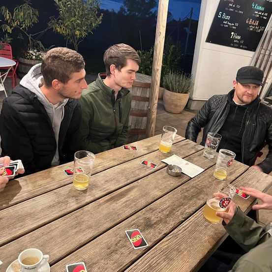Vier Männer sitzen um einen Holztisch in einer Kneipe, halten Gläser mit Bier in den Händen und spielen ein Kartenspiel.