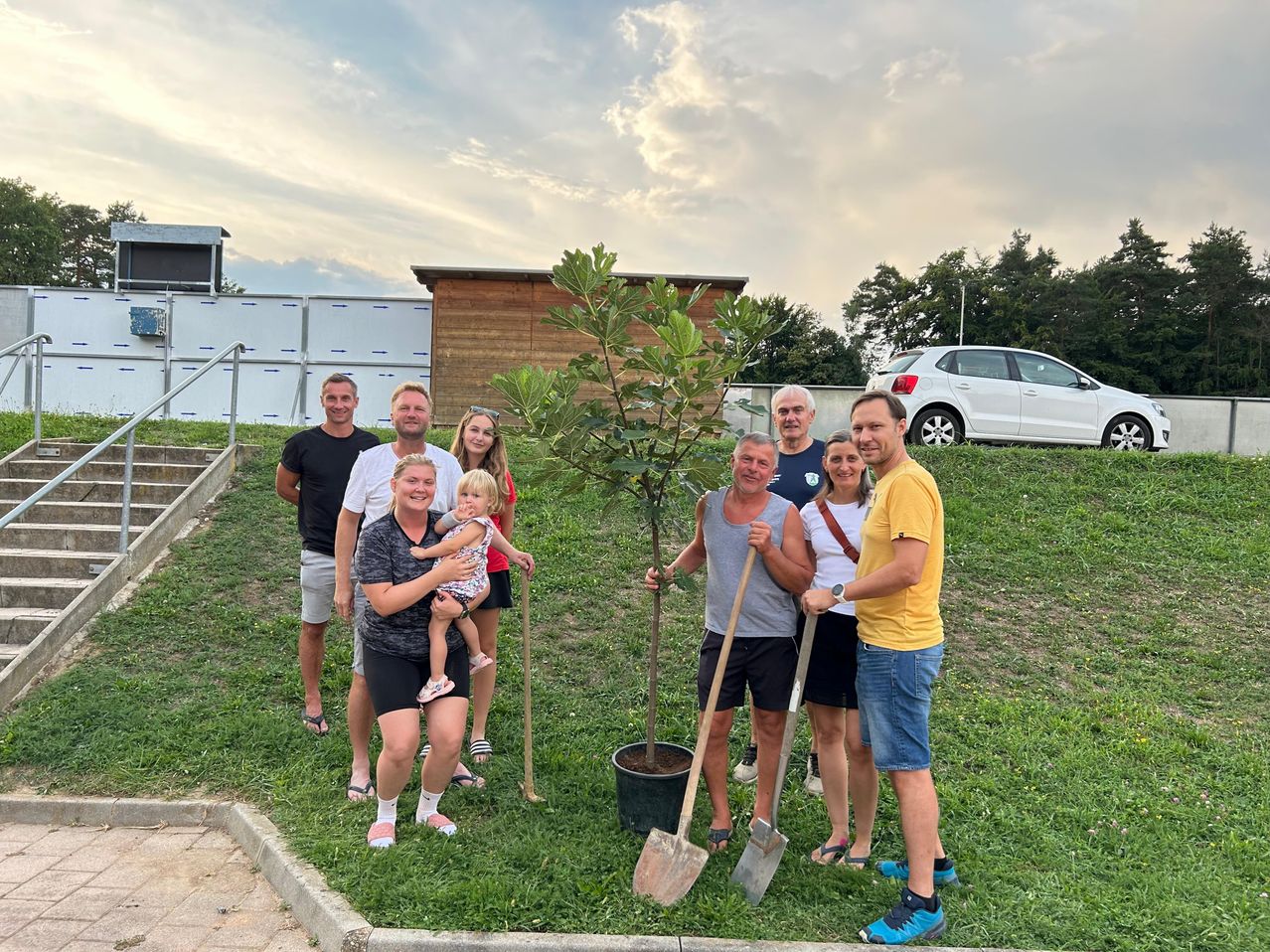 Eine Gruppe von Menschen posiert für ein Foto in einem Park. Sie stehen um einen kleinen Baum herum und halten Schaufeln. Eine Frau hält ein Baby, und ein Auto ist in der Ferne geparkt.
