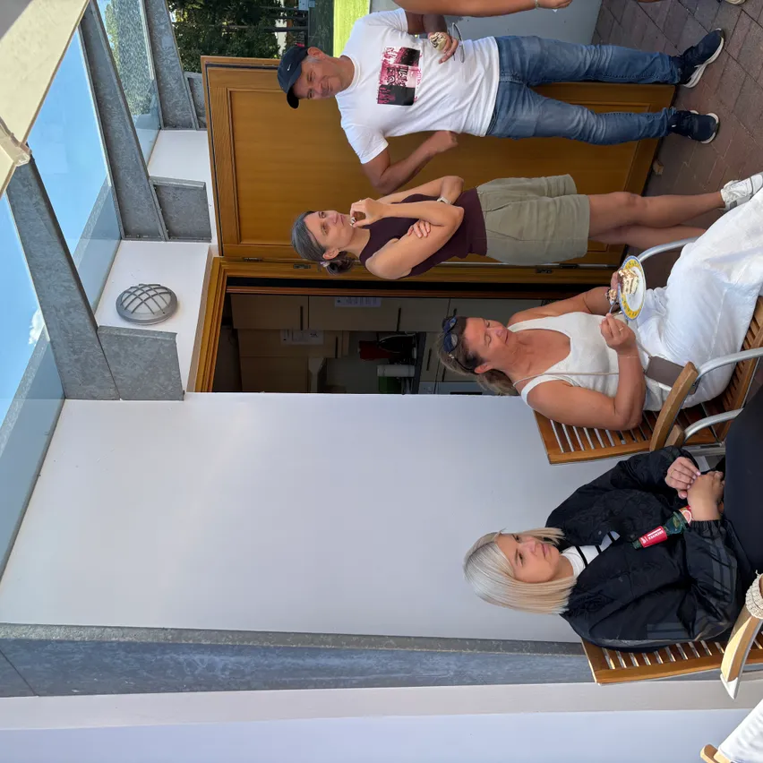 Eine Gruppe von Menschen versammelt sich auf einer Terrasse. Eine Frau sitzt auf einem Stuhl und isst einen Nachtisch. Eine andere Frau lehnt sich an den Türrahmen. Ein Mann steht hinter ihnen.
