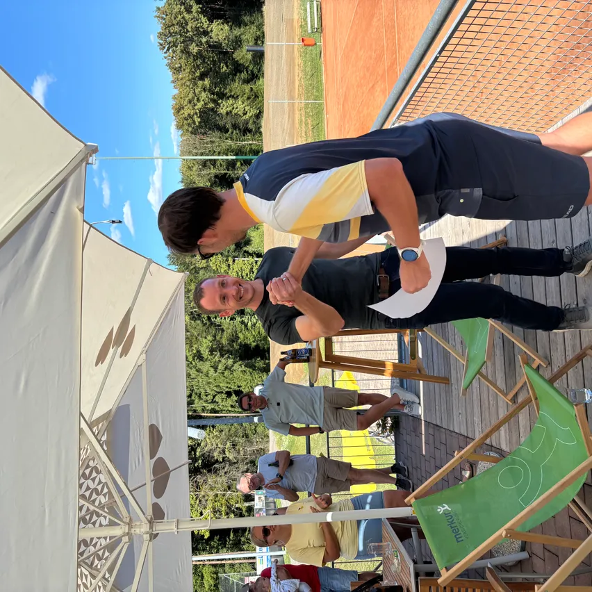An einem sonnigen Tag versammeln sich mehrere Personen auf einer Holzterrasse, wobei eine Person ein Stück Papier in der Hand hält. Dahinter befindet sich ein Baseballfeld.