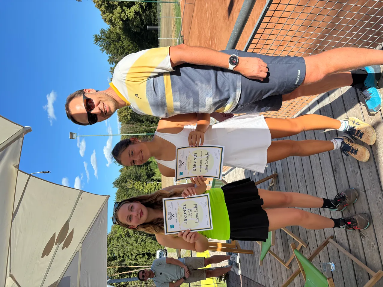 Drei Personen posieren auf einem Tennisplatz für ein Foto, zwei halten Urkunden. Eine Frau trägt ein grünes Oberteil, die andere ein weißes Tanktop. Ein Mann steht hinter ihnen und lächelt.