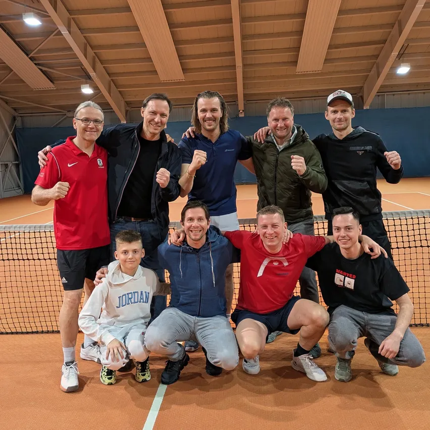 Eine Gruppe von Männern posiert für ein Foto auf einem Tennisplatz. Sie tragen verschiedenfarbige Hemden und Hosen. Sie scheinen für ein Teamfoto zu posieren.