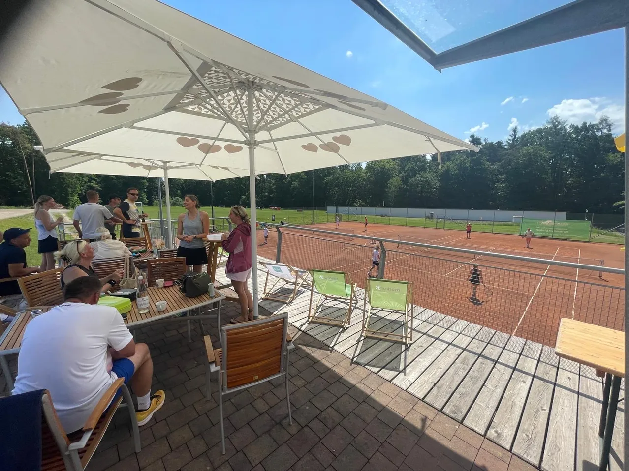 Eine Gruppe von Menschen sitzt unter einem großen Schirm auf einer Terrasse. Im Hintergrund üben Tennisspieler auf einem von einem Zaun umgebenen Platz. Der Himmel ist klar mit ein paar Wolken.