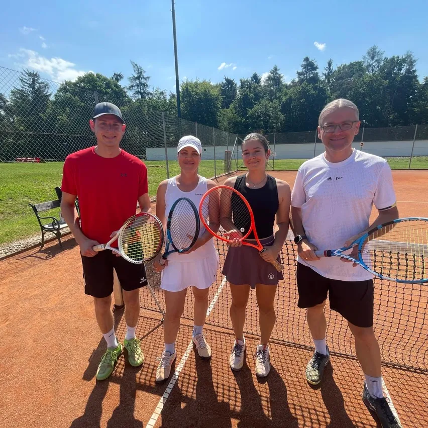 Vier Personen stehen auf einem Tennisplatz, zwei Frauen in der Mitte, halten Tennisschläger, und zwei Männer an den Seiten, alle lächeln für die Kamera.
