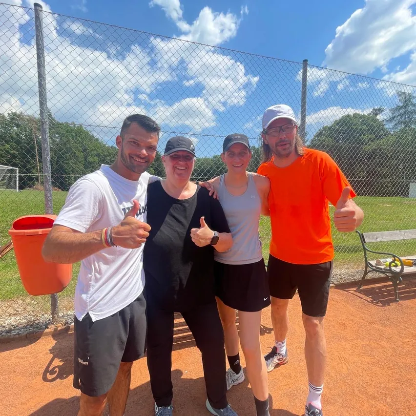 Vier Personen stehen auf einem Sportplatz und posieren für ein Foto. Ein Mann zeigt den Daumen nach oben, während zwei Frauen und ein weiterer Mann lächeln. Im Hintergrund befinden sich ein Zaun, Bäume und eine Bank.