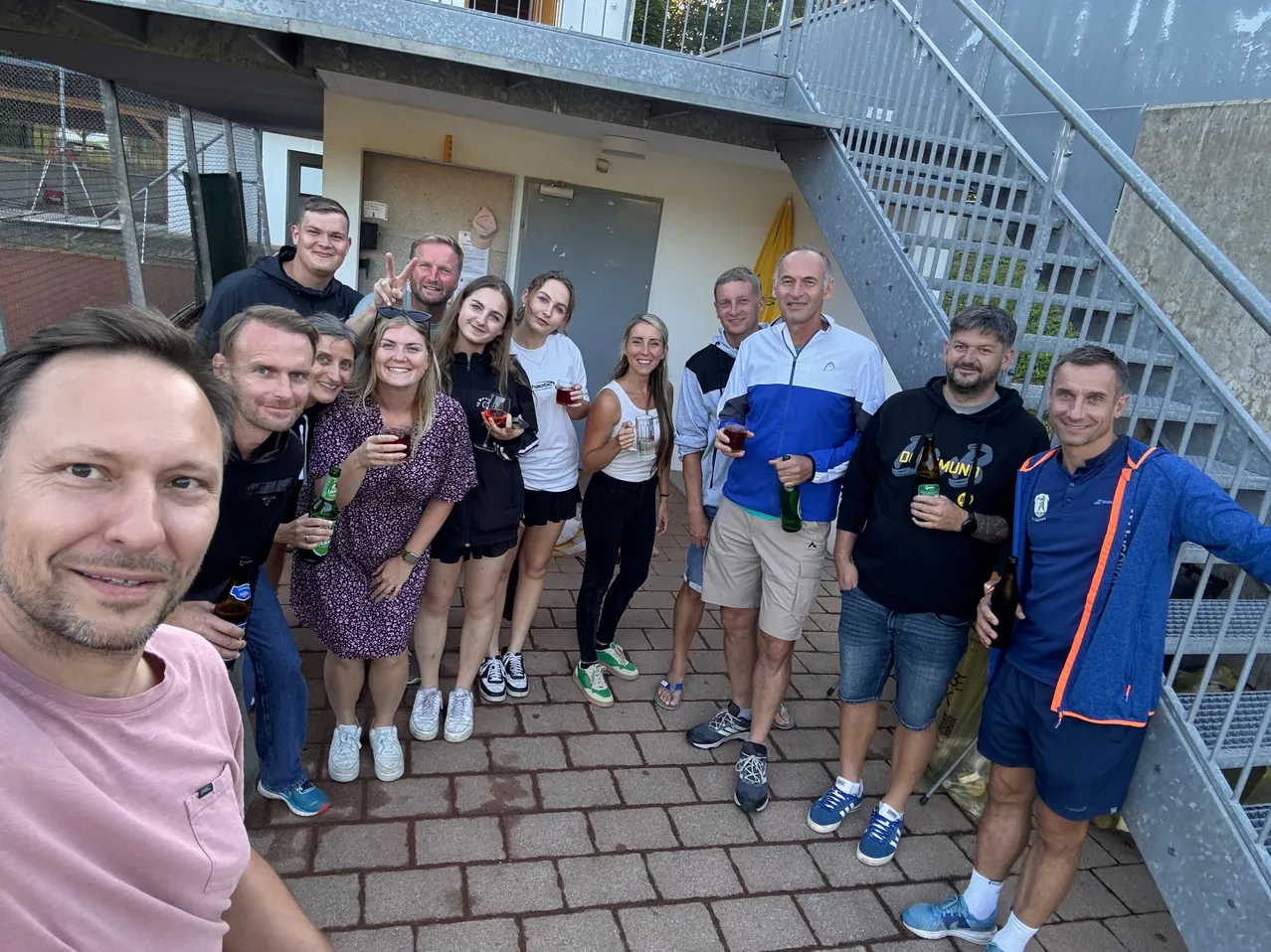 Eine Gruppe von Männern und Frauen steht vor einem Gebäude, lächelt und hält Getränke in der Hand. Sie sind lässig gekleidet und scheinen für ein Foto zu posieren.