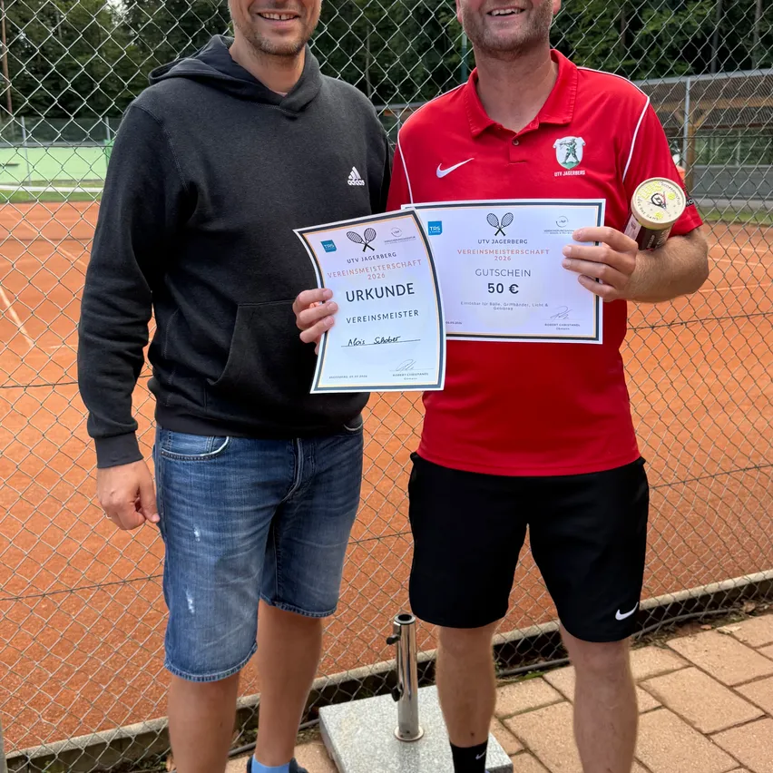 Zwei Männer stehen auf einem Tennisplatz, einer hält ein Zertifikat und einen Pokal, der andere ein Zertifikat. Sie lächeln und posieren für ein Foto.