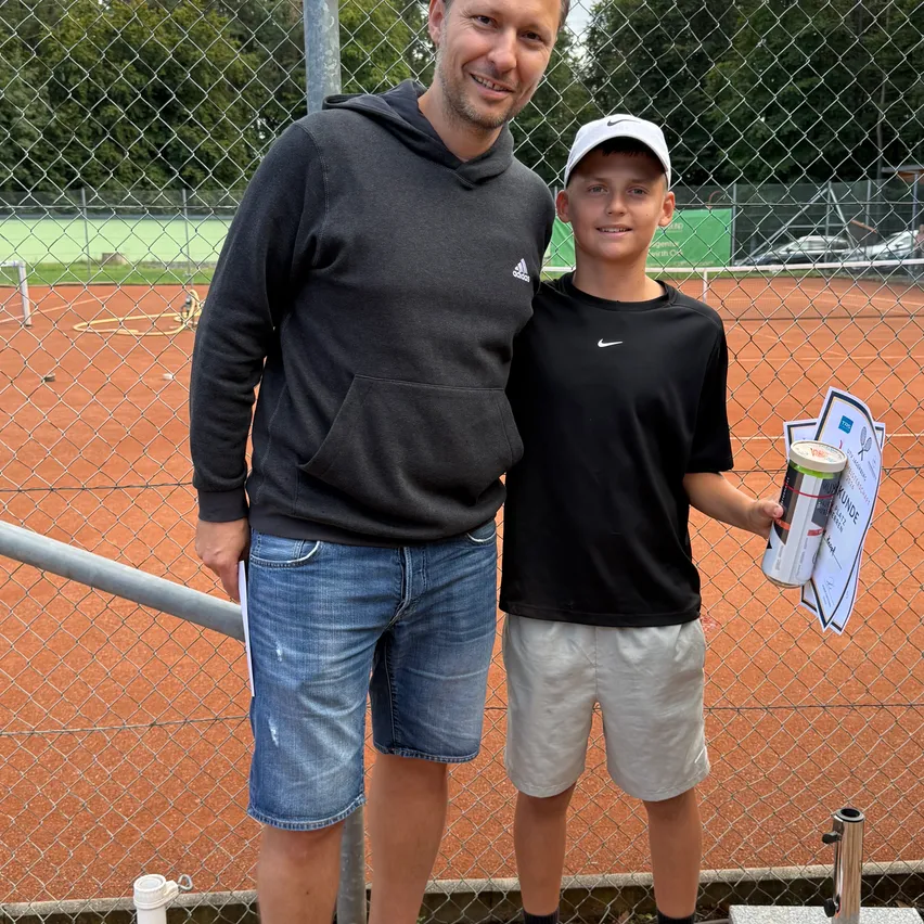 Ein Mann und ein Junge stehen vor einem Tennisplatzzaun und lächeln. Der Junge hält einen Pokal und ein Zertifikat.